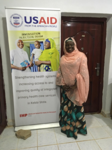WI-HER'S Kebbi State Gender, Social Inclusion and Community Engagement Advisor, Nafisa Zaki, poses next to the USAID Integrated Health Program Banner.